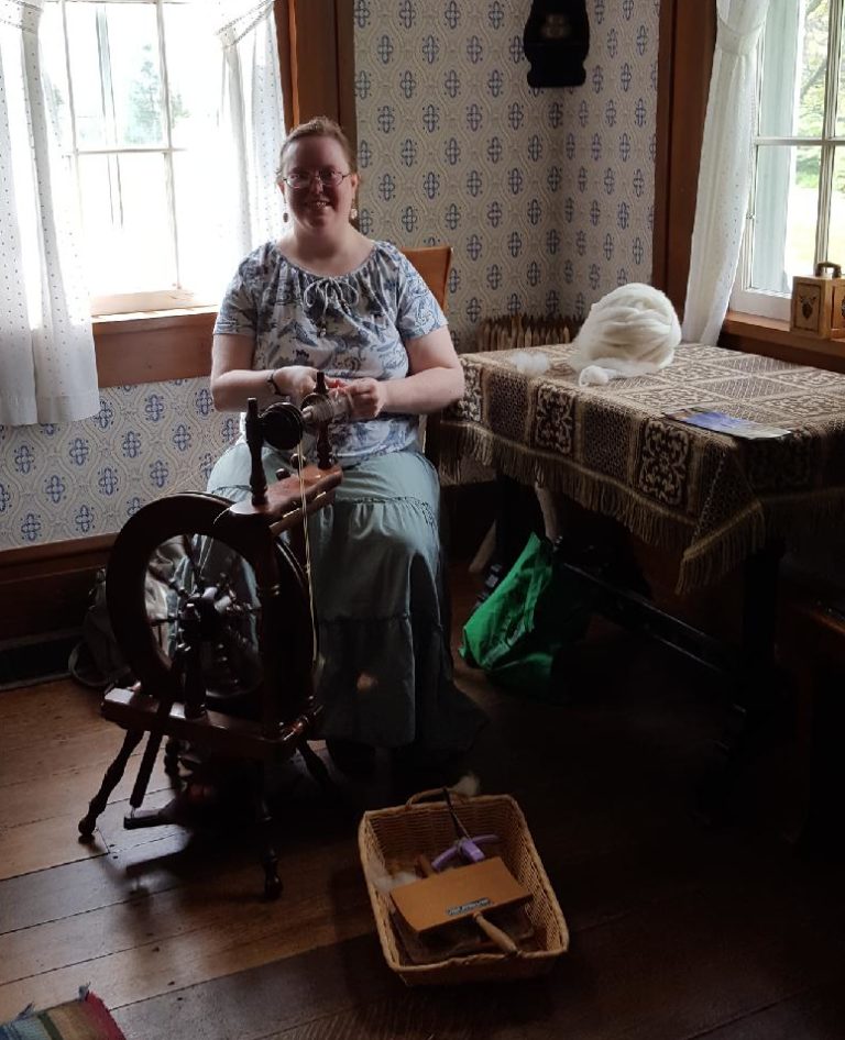 A smiling blonde woman with glasses sits at a spinning wheel in the corner of an antique farm house